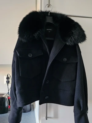 Svart pälsjacka från Meotine - Snygg svart pälsjacka från Meotine med stor fluffig fuskpälskrage. Jackan har två stora bröstfickor med lock och knappar framtill. Perfekt för kalla vinterdagar och ger en cool och lyxig vibe till din outfit.