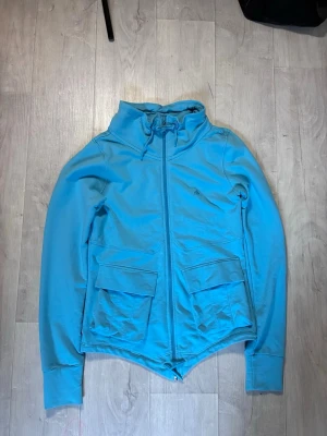 Blå Adidas vindjacka med dragkedja - Snygg blå Adidas vindjacka med hög krage och hel dragkedja framtill. Jackan har två stora fickor med lock och snörning i halsen. Perfekt för dig som vill ha en sportig och stilren look.