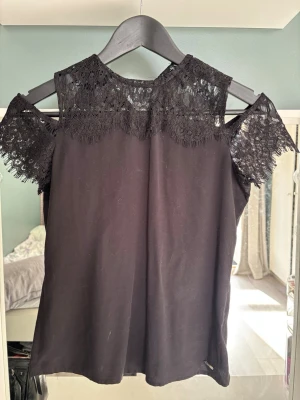 Svart spetsdetaljerad topp  - Snygg svart topp från Guess med spetsdetaljer över axlar och halsringning. Toppen har korta ärmar med öppen axel och en feminin, elegant look. Perfekt för dig som gillar stilrena plagg med det lilla extra.