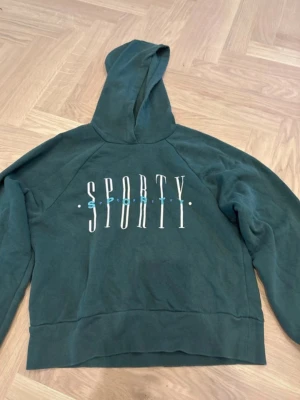 Grön hoodie med SPORTY-tryck - Mörkgrön hoodie med huva och stort vitt tryck där det står SPORTY på bröstet. Hoodien har en avslappnad passform och ribbade muddar vid ärmslut och nederkant. Perfekt för en chill och sportig look.