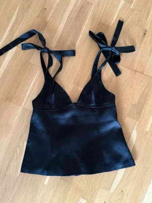 Svart satinlinnetopp - Superfin topp i satin med rosettdetaljer på axelbanden. Aldrig använd då den tyvärr är lite för liten för mig. 
