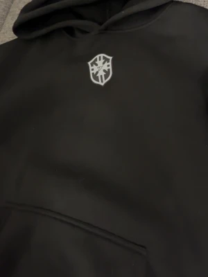 Svart hoodie med vit brazil logga - Svart hoodie med en vit logga på bröstet. Klassisk modell med huva och stor magficka framtill. Perfekt för en chill och avslappnad stil. Materialet ser ut att vara mjukt och bekvämt, passar bra till jeans eller joggers.