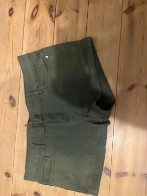 Mörkgröna jeansshorts med fickor - Snygga mörkgröna shorts i jeansmodell med klassiska fem fickor och knappstängning. Shortsen har dekorativa sömmar på bakfickorna och är perfekta för varma dagar. Passar dig som gillar en enkel och stilren look. Iandvånda