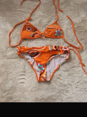 Orange blommig bikini med knyt - Superfin orange bikini med blommigt mönster och retrovibbar. Överdelen har trekantsform med knyt i nacken och ryggen, och underdelen har knyt i sidorna. Materialet är stretchigt och bekvämt, perfekt för stranden eller poolen. Passar s