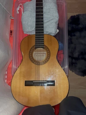 Klassisk gitarr från Cataluña - Säljer en klassisk akustisk gitarr från Cataluña, modell SGN-C61. Gitarren har en naturlig träfärg med mörkare detaljer runt ljudhålet och svart greppbräda. Perfekt för dig som vill börja spela eller bara jamma hemma.