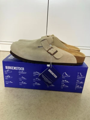 Beige Birkenstock Boston tofflor mocka - Säljer ett par snygga beige Birkenstock Boston tofflor i mjuk mocka. De har en justerbar rem med metallspänne och klassisk korksula för extra komfort. Perfekta för chill dagar hemma eller när du vill ha en avslappnad stil.