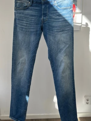 Blå slim fit jeans från Jack & Jones W32/L34 - Snygga blå slim fit jeans från Jack & Jones, modell Glenn. Jeansen har klassisk femficksdesign, normal midja och lätt tvättad look med subtila slitningar. Perfekta för en trendig och avslappnad stil.