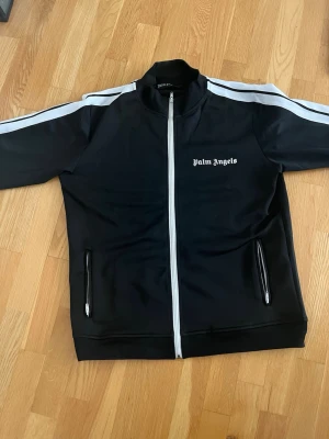 Svart track jacket från Palm Angels - Svart track jacket från Palm Angels med vita ränder längs ärmarna och vit dragkedja framtill. Storlek large, pris kan diskuteras.