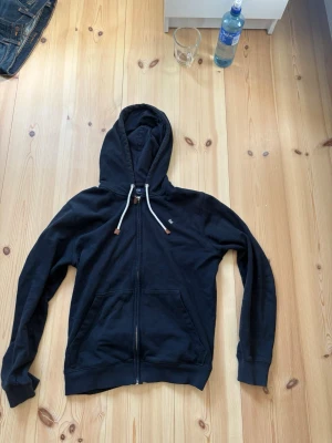 Mörkblå hoodie från Morris  - Mörk blå hoodie från Morris med dragkedja framtill, vita snören i huvan och två fickor. Klassisk och stilren design med ribbade muddar och liten logga på bröstet. Perfekt för en avslappnad look.