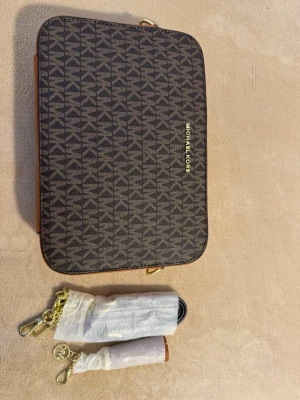 Brun axelväska från Michael Kors - Snygg brun axelväska från Michael Kors med guldiga detaljer och logomönster över hela väskan. Rektangulär form och justerbar axelrem i kedja och läder. Perfekt för dig som gillar stilrena accessoarer med lyxig känsla.
