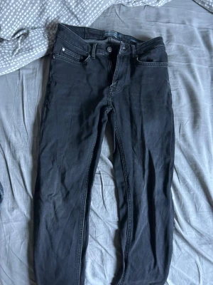 Svarta slim fit jeans från Dressmann - Svarta jeans från Dressmann i slim fit-modell. Klassisk femficksdesign med dragkedjegylf och bälteshällor. Jeansen är tillverkade i ett stretchigt material för en bekväm passform och har diskreta sömmar i matchande färg.
