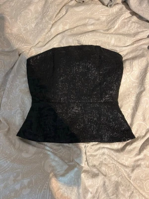 Svart glittrig korsettopp H&M - Svart glittrig korsettopp från H&M med figurnära passform. Toppen är axelbandslös och har en lätt utsvängd nederkant. Perfekt för dig som vill sticka ut med en snygg och trendig look.