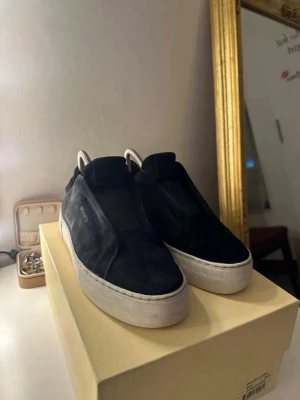  Arigatos  - Stilrena svarta sneakers från Axel Arigato med ovandel i mocka och diskret logga på sidan. Skorna har en tjock vit platt sula och minimalistisk design utan synlig snörning. Perfekta för en clean och modern look. Allt medföljer Låda dustbag 