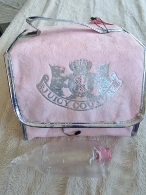 Juicy Couture rosa necessärväska - Snygg rosa necessärväska från Juicy Couture med silverdetaljer och logga framtill. Väskan har flera genomskinliga fack inuti och ett praktiskt handtag upptill. Perfekt för att organisera smink eller hudvård när du är på språng.
