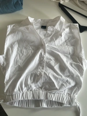 Vit ärmlös blus med krage från Gina Tricot - Stilren vit blus från Gina Tricot med klassisk krage och knäppning framtill. Blusen är ärmlös och har en elastisk dragsko i midjan för en snygg passform. Perfekt att matcha med jeans eller kjol för en fräsch look.