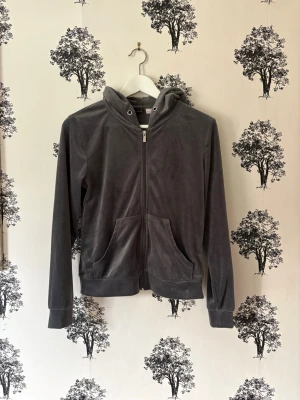 Mörkgrå velourhoodie med dragkedja - Mjuk och skön mörkgrå hoodie i velour med dragkedja framtill och två stora fickor. Tröjan har huva och ribbade muddar vid ärmslut och nederkant. Perfekt för en avslappnad och trendig look.