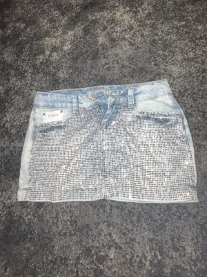 Ljusblå jeanskjol med paljetter - Snygg ljusblå jeanskjol från Philosophy Blues Original med glittriga paljetter framtill. Klassisk femficksmodell och kort passform. Perfekt för dig som vill sticka ut med en unik denimlook.