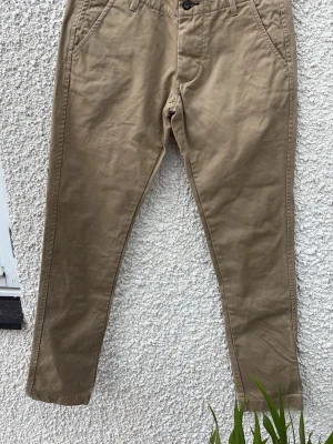 Beiga chinos från NN07 - Snygga beiga chinos från NN07 med klassisk rak passform och två sidofickor. Byxorna har en stilren design med knappstängning och detaljerad insida i midjan med randigt tyg. Perfekta för en avslappnad men ändå uppklädd look.