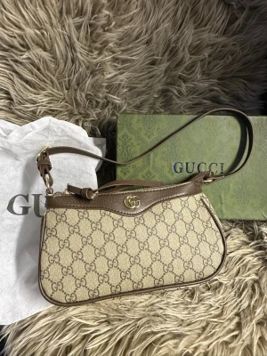 Beige axelremsväska  - Säljer en beige axelremsväska med klassiskt GG-monogram i canvas och bruna läderdetaljer. Väskan har guldfärgade metalldetaljer och dragkedja upptill. Perfekt storlek för det viktigaste och en ikonisk accessoar för din outfit.