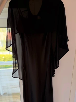 Svart långklänning med transparent sjal - Svart långklänning med smala axelband och rak siluett. Till klänningen hör en transparent, vid sjal som ger en elegant look. Perfekt för dig som vill ha en stilren och klassisk outfit. Materialet känns lätt och följsamt.