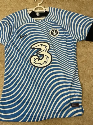 Chelsea FC matchtröja blåvit Nike - Chelsea FC matchtröja i blått och vitt med vågiga ränder, officiell klubbmärke på bröstet och Nike-logga. Tillverkad i lätt och ventilerande material, perfekt för fotbollsträning eller match. Rund halsringning och korta ärmar.