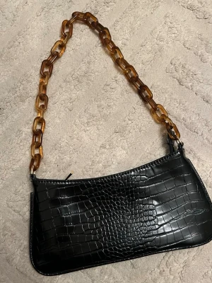 Svart croco handväska med kedja - Snygg svart handväska i croco-mönstrat konstläder med en chunky kedjerem i brunmelerad plast. Väskan har en avlång form och stängs med dragkedja upptill. Perfekt accessoar för att lyfta din outfit.