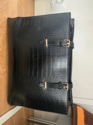 Svart handväska i krokopräglat material - Stilren svart handväska i krokopräglat konstläder med två dekorativa spännen i guld och tillhörande nyckelfodral. Väskan har en fyrkantig form och rymmer mycket, perfekt för dig som gillar att bära med dig allt du behöver. Snygg och trendig accessoar till din outfit.