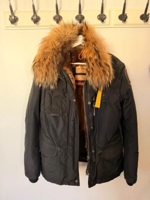 Svart parkas från Parajumpers med päls - Snygg svart parkas från Parajumpers med stor, fluffig päls runt kragen och flera praktiska fickor. Jackan har dragkedja, knappar och den klassiska gula Parajumpers-detaljen. Perfekt för kalla dagar när du vill hålla stilen. Äkta jacka och äkta päls. Finns luva som man kan plocka ut och stoppa in ✨