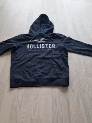 Svart Hollister hoodie med logga - Snygg svart hoodie från Hollister med stor vit logga och texten 'California' på bröstet. Klassisk känguruficka framtill och justerbar huva. Tillverkad i mjukt bomullsmaterial som är skönt att ha på sig. Perfekt för en avslappnad och cool stil.