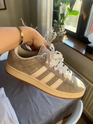 Adidas Campus grå sneakers - Grå Adidas Campus sneakers i mocka med vita detaljer och klassiska tre ränder på sidorna. Skorna har vit sula och vita skosnören, samt Adidas-logga på plösen. Skorna är i mycket bra skicka 