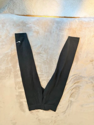 Svarta leggings från Nike - Säljer ett par svarta leggings från Nike med klassisk vit swoosh-detalj på ena benet. De har hög midja och är tillverkade i ett stretchigt material som sitter skönt och tight. Perfekta för träning eller chill dagar.