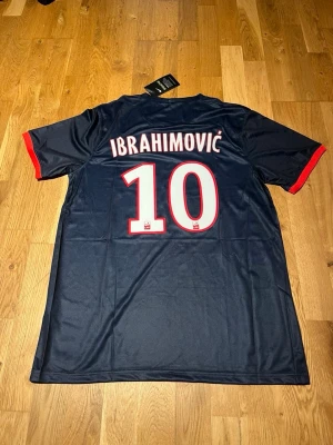 PSG Ibrahimović matchtröja Nike - Mörkblå Paris Saint-Germain matchtröja med röd mittremsa, vita detaljer och röda ärmslut. Tryck med Ibrahimović och nummer 10 på ryggen. Nike-logga och Fly Emirates på bröstet. Tillverkad i lätt och ventilerande Dri-FIT-material.