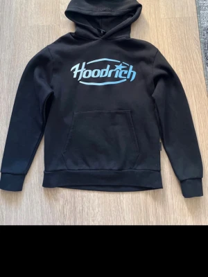 Svart hoodie från Hoodrich XS - Svart hoodie från Hoodrich med stor blå logga på bröstet och klassisk känguruficka framtill. Mjuk insida och ribbade muddar vid ärmslut och nederkant. Perfekt för en chill och streetinspirerad stil.