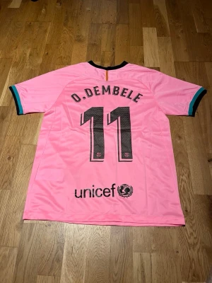 Barcelona third kit 20/21 O.dembele #11 - Snygg rosa FC Barcelona matchtröja från Nike med O. Dembele och nummer 11 på ryggen. Tröjan har svarta och turkosa detaljer vid ärmslut och krage, samt klubbmärke och sponsortryck på bröstet. Perfekt för fotbollsfans!