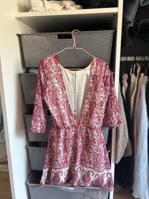 Rödvit mönstrad kortklänning - Superfin kortklänning med rödvitt blommigt mönster och öppen rygg med en elegant och snygg kedja som hänger superfint längs ryggen. Klänningen har trekvartsärm och markerad midja, vilket ger en snygg siluett. Perfekt för sommarens alla tillfällen. Materialet känns lätt och luftigt.☀️🌸 Pris kan absolut diskuteras!❣️