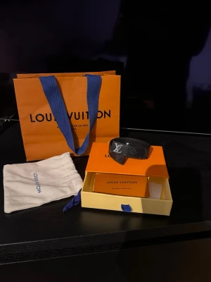 LOUlS VUITTON svart armband  - Snyggt svart armband med klassiskt monogrammönster och en silverfärgad logga framtill. Tillverkat i läder och levereras med original ask, dustbag och påse. Perfekt accessoar för dig som gillar exklusiv stil. Passar dig som har en handled som är 17 - 18,5 - 20cm runt handleden. Storleken är 21. Lite slitningar kan skicka bild med det är inget man ser eller tänker på 