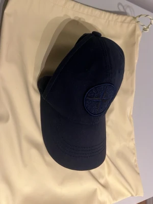 Mörkblå keps från Stone Island - Snygg mörkblå keps från Stone Island med broderad logotyp framtill i samma färg som kepsen. Justerbar rem baktill i metall och klassisk böjd skärm. Tillverkad i bomull för en bekväm och stilren look.