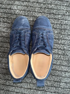 Blåa sneakers från Christian Louboutin - Snygga blå sneakers i mocka från Christian Louboutin med klassisk röd sula och diskreta sömdetaljer. Skorna har lågt skaft, rund tå och matchande blå skosnören. Insidan är ljusbeige och sulan har den ikoniska Louboutin-signaturen.