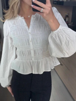 Vit blus med volang och puffärm - Söt vit blus med knappar framtill, smockad överdel och volang nertill. Blusen har långa puffärmar med resår vid ärmslut och lätt rynkad struktur. Perfekt för en trendig och feminin look.