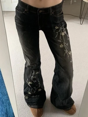 Svarta jeans med blommig brodyr - Snygga svarta jeans med utsvängda ben och broderade blommor i vitt och beige längs ena benet. Jeansen har klassisk femficksmodell och hög midja. Perfekta för dig som vill sticka ut med en unik detalj.
