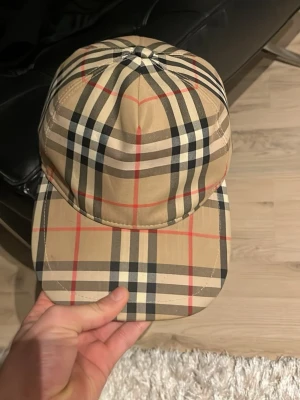 Beige Burberry keps  - Säljer en klassisk beige keps från Burberry med det ikoniska rutmönstret i svart, vitt och rött. Kepsen har böjd skärm och justerbar snapback i svart plast. Märkt med Burberry London England baktill. Perfekt för dig sommar keps 