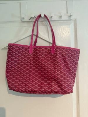 Rosa väska från Goyard - Snygg rosa väska från Goyard med klassiskt mönster i vitt och rosa. Väskan har dubbla handtag och är rymlig med mjuk form, perfekt för att bära med sig allt man behöver. Tillverkad i canvas med detaljer i läder.
