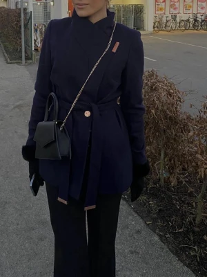 Mörkblå Ted Baker-kappa med skärp - Stilren mörkblå kappa med asymmetrisk knäppning och matchande midjeskärp. Kappan har en dekorativ knapp framtill och bred krage som kan bäras både öppen och stängd (dold magnet som gör att den kan vara stängd). Perfekt för dig som gillar minimalistisk design och vill ha en snygg jacka till hösten. Mycket bra skick, nypris ca 4749 kr.