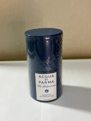 Acqua di Parma Blu Mediterraneo parfym - Acqua di Parma Blu Mediterraneo Ginepro di Sardegna är en elegant parfym i en djupblå cylinderformad förpackning. Märket är känt för sina exklusiva dofter och denna variant har en fräsch, medelhavsinspirerad känsla. Förpackningen är av plast och kartong med vit etikett och blå text.