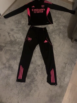 Arsenal träningsset svart/rosa Adidas - Säljer ett Arsenal träningsset från Adidas, bestående av långärmad tröja och matchande byxor. Setet är svart med rosa detaljer och loggor, tillverkat i ett lätt och funktionellt material som passar perfekt för fotbollsträning eller gym. Tröjan har tryck med Emirates och klubbmärke.