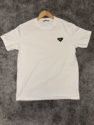 Vit t-shirt från Prada - Snygg vit t-shirt från Prada med klassisk rund halsringning och diskret svart logotyp-patch på bröstet. Enkel och stilren design som passar till det mesta. Tillverkad i mjuk bomull för en skön känsla.