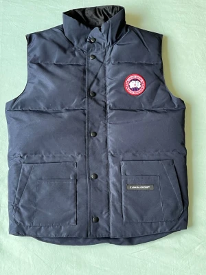 Svart dunväst från Canada Goose - Stilren svart dunväst från Canada Goose med hög krage, två stora fickor framtill och klassisk logotyp på bröstet. Västen har tryckknappar och är perfekt för lager-på-lager-stil under kyliga dagar.