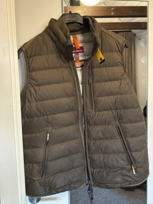 Mörk grön dunväst från Parajumpers - Snygg militär grön dunväst från Parajumpers med quiltad design och hög krage. Västen har två dragkedjefickor framtill och en gul detalj vid kragen. Superlätt och perfekt för lager-på-lager-stil.