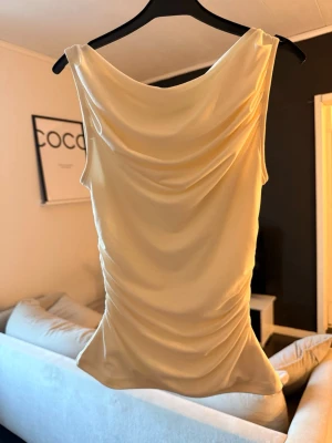 Crémevit omlottopp i satin - Snygg crémevit topp i satin med draperad framsida och omlottkänsla. Toppen är ärmlös och har en mjuk, glansig finish som ger en lyxig vibe. Perfekt att styla med både jeans och kjol för en trendig look.