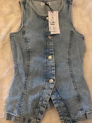 Ljusblå ärmlös jeanslinnetopp XS - Ljusblå ärmlös linnetopp från Gina Tricot i denim. Toppen har knappar framtill, figurnära passform och en liten slits nedtill. Perfekt för dig som vill ha en trendig och unik topp i garderoben.